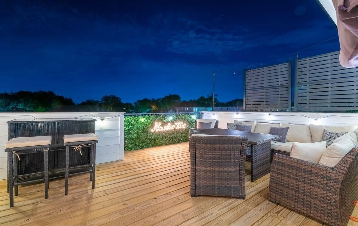 Um pátio externo com móveis de vime, um letreiro neon e vista para o horizonte da cidade ao entardecer.
