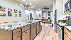 A modern kitchen with a granite countertop, wooden cabinets, and stainless steel appliances. There is a dining area with a brown leather couch and a small rug. - Huge Downtown Luxury Home Rooftop Deck 12 Beds (Nashville)