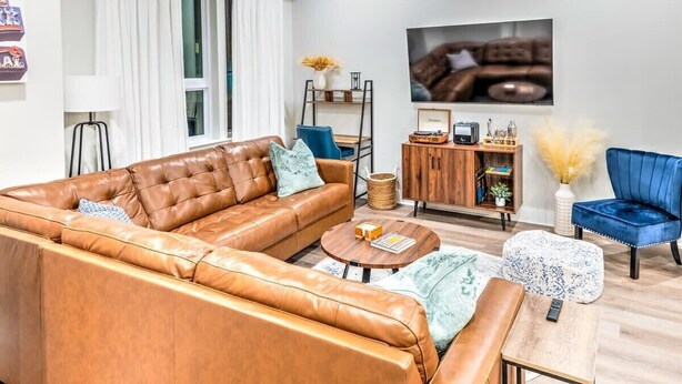 A modern living room with a brown leather sofa, a blue armchair, a wooden coffee table, and a TV mounted on the wall. - Huge Downtown Luxury Home Rooftop Deck 12 Beds (Nashville)