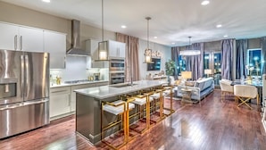 A modern kitchen with stainless steel appliances, a central island with bar stools, and a dining area with a sofa and chandelier. - Rooftop Pool Table Gulch! City Views! 15 beds! (Nashville)