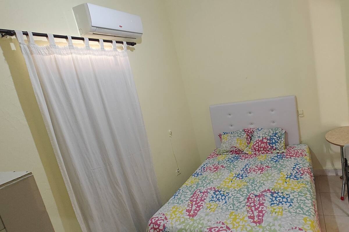 A small, single-bed room with a colorful bedspread, a white curtain, a small stool, and an air conditioning unit.