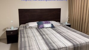 A neatly made bed with a striped bedspread and two decorative pillows, a bedside table with a lamp, and a curtain on the window. - Surbackpackers (Santa Cruz)