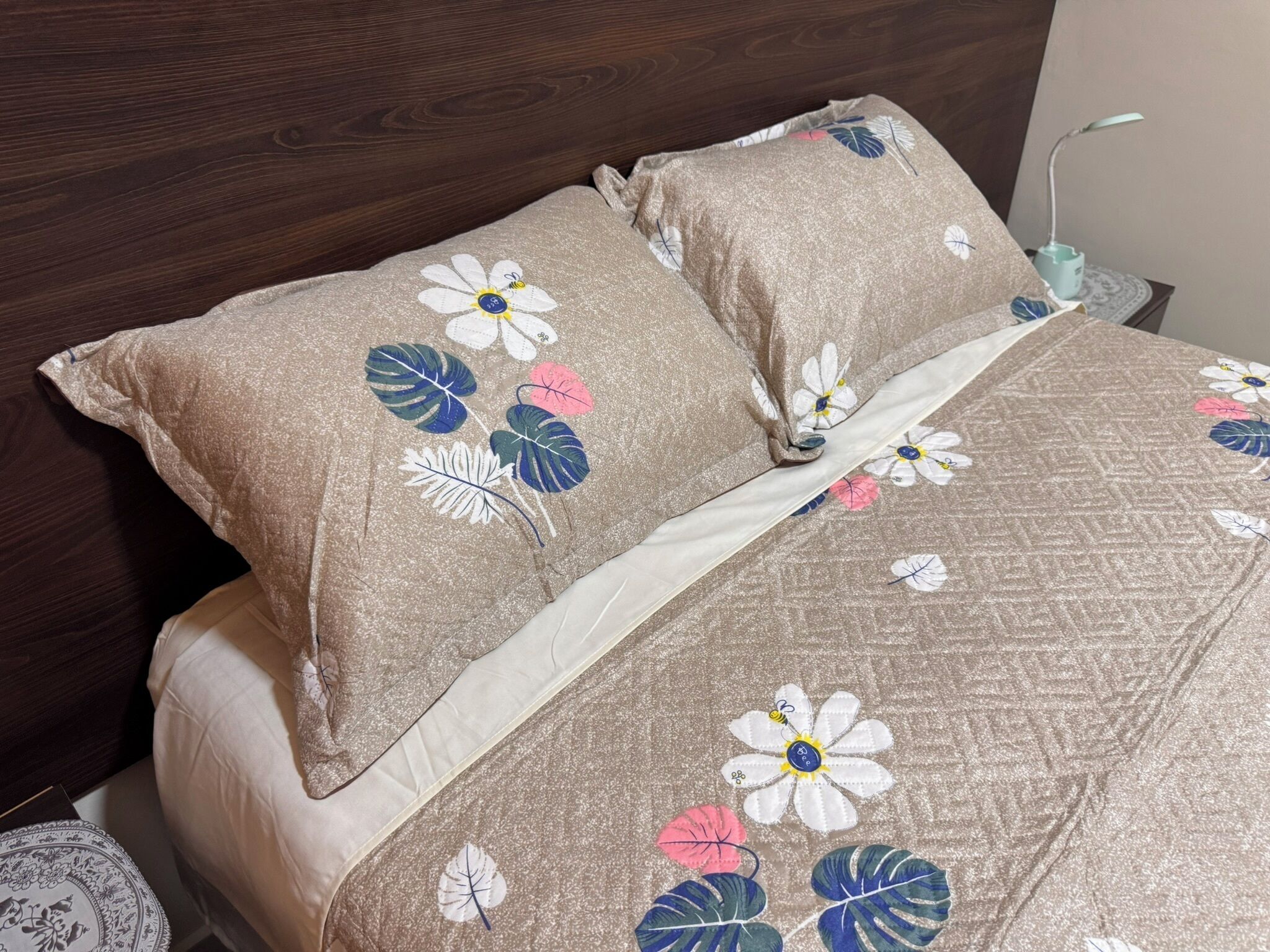 Ein Bett mit blumigem Bettbezug und Kissen, ein hölzernes Kopfteil und ein Nachttischchen mit Lampe.