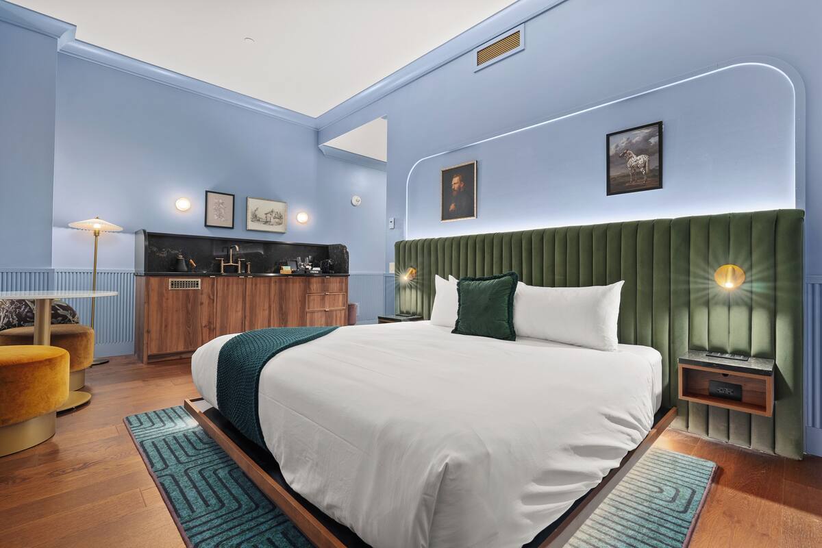 Quarto com cama grande, cabeceira estofada verde, mesa de cabeceira com abajur e cômoda de madeira pequena.