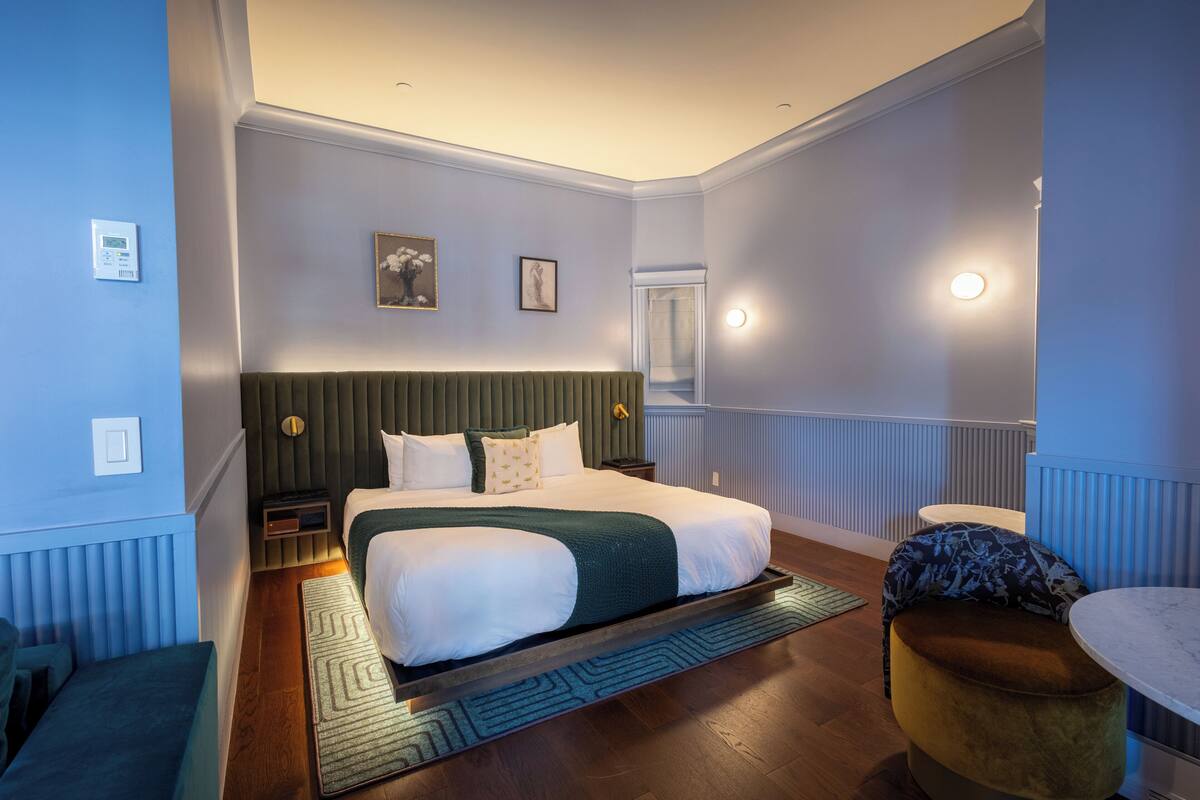 Quarto de hotel moderno com uma cama grande, cabeceira verde, mesa de cabeceira pequena, criado-mudo e um banco.
