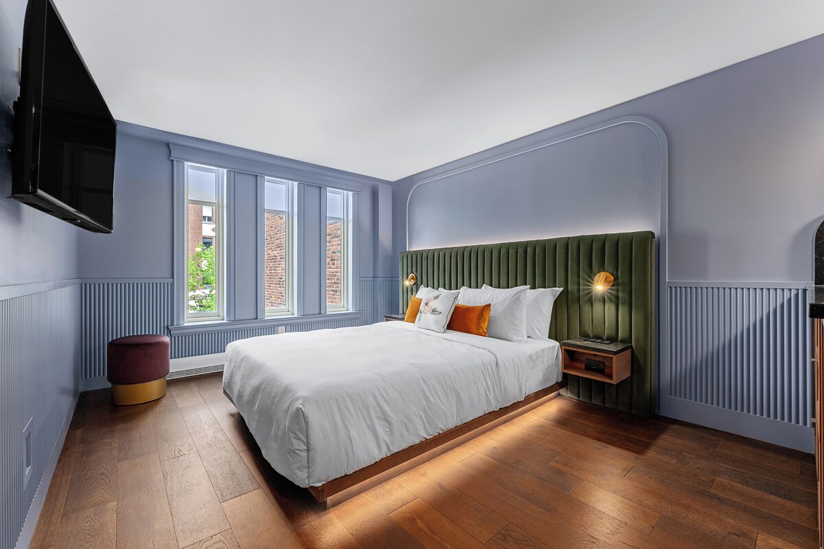 A bedroom with a bed, nightstands, a TV, and a window with a view of a brick building.