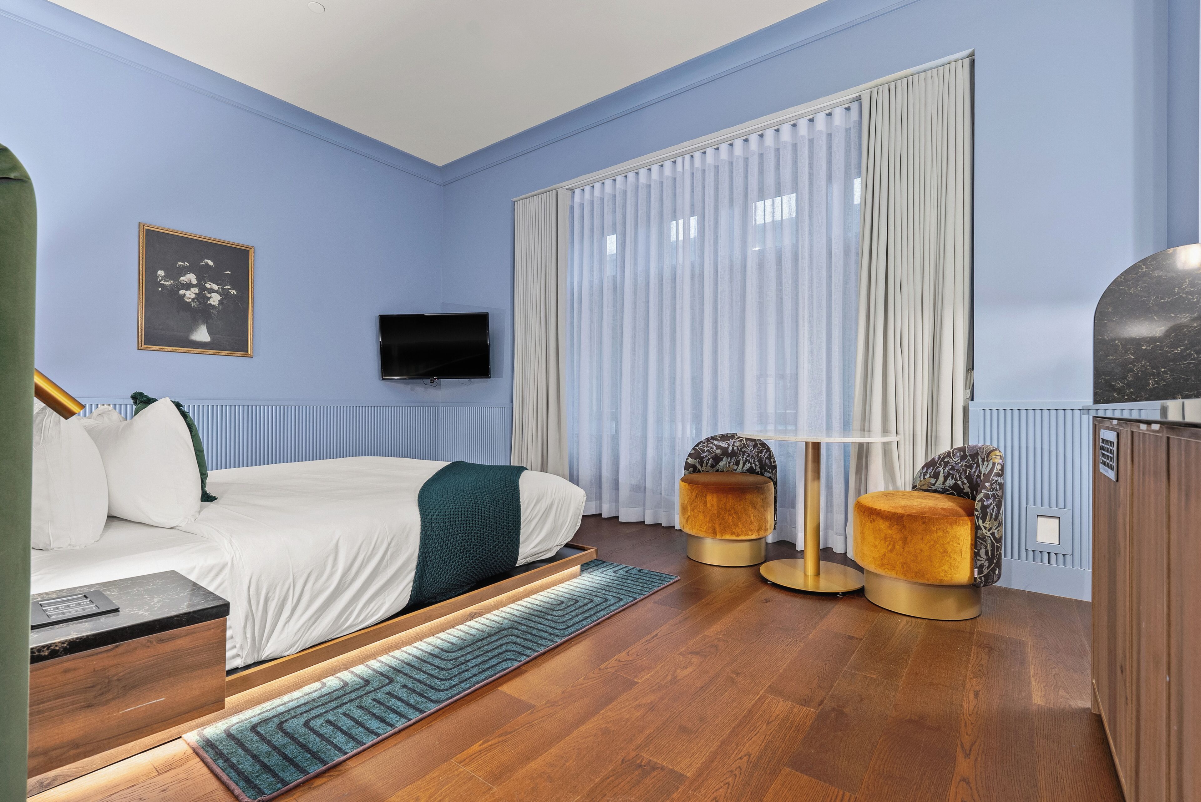 Un dormitorio moderno con cama, televisor, ventana con cortinas y un pequeño rincón de descanso con mesa y sillas.