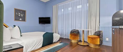 Un dormitorio moderno con cama, televisor, ventana con cortinas y un pequeño rincón de descanso con mesa y sillas.