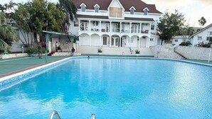 A swimming pool with a clear blue water, surrounded by a white building with a red roof and multiple balconies.