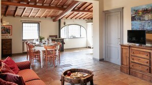A spacious living area with a high wooden ceiling, a dining table set for four, a sofa, and a television on a wooden cabinet. - Villa Peramonda (Toscana)