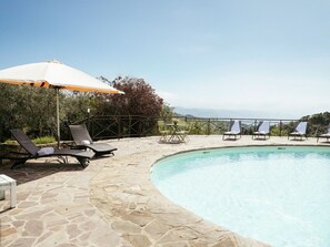 Un espace aménagé au bord de la piscine, avec des chaises longues et un parasol, entouré d’un sol en pierre et offrant une vue sur le paysage.