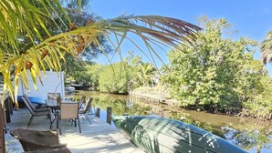 A wooden deck with wicker chairs, a table, and a green kayak by a calm water body, surrounded by lush greenery. - Private Waterfront Oasis w/Heated Pool/Hot Tub & Dock, Close to Everything! (Naples)