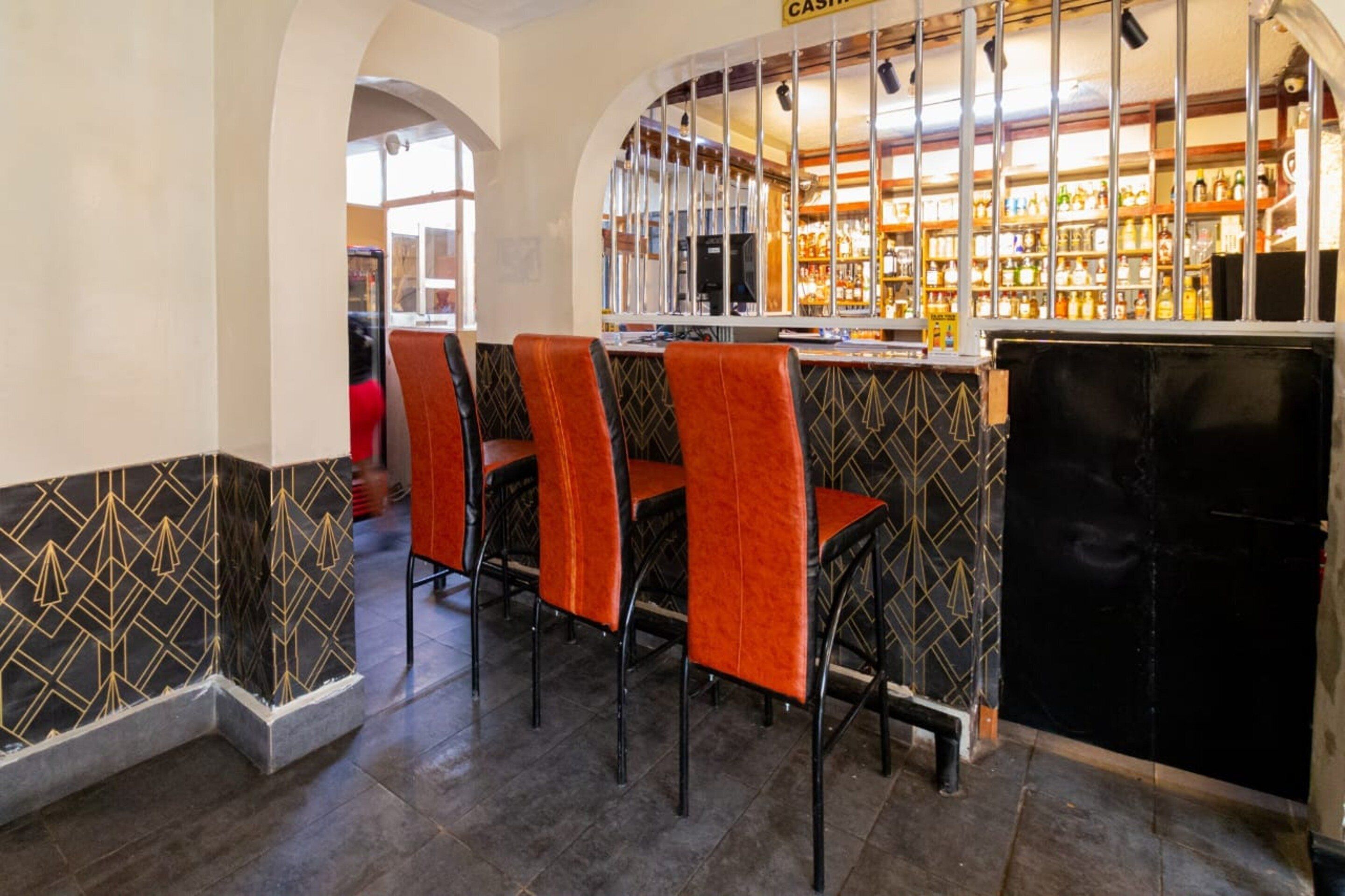 Angolo bar con bancone, cassa e selezione di bottiglie. Quattro sedie con rivestimento arancione.