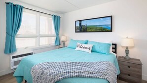 Un dormitorio con cama, ventana con cortinas, un cuadro en la pared y mesita de noche con lámpara.
