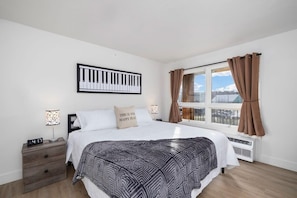 Ein Schlafzimmer mit Bett, Nachttisch, Wanduhr, einem Fenster mit Vorhängen und Blick nach draußen.