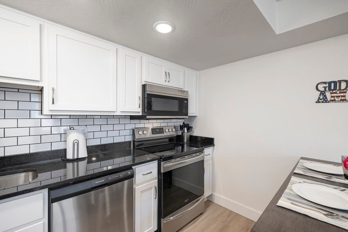 A compact kitchen with white cabinets, a black countertop, stainless steel appliances, and a dining area with a table set for two.
