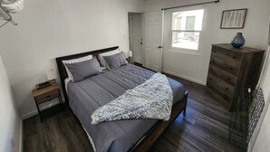 Een slaapkamer met een bed, een kaptafel, een nachtkastje, een raam en een deur.