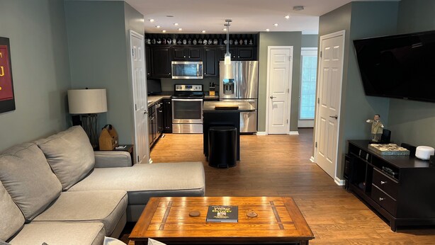 Una sala de estar moderna con un sofá, una mesa de centro y un televisor. Se ve una cocina con electrodomésticos y un comedor al fondo.