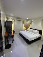 Quarto de hotel com uma cama, uma escrivaninha com cadeira, ar condicionado e cortinas.
