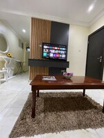 Uma sala de estar com uma TV de tela plana na parede, uma mesa de centro de madeira com uma revista e um tapete felpudo.