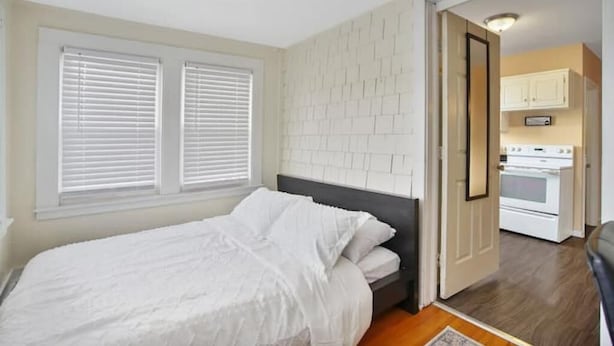 A bedroom with a bed, white blinds, a white brick wall, and a kitchen visible through an open door.