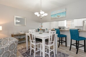 Un coin repas avec une table et des chaises blanches, un lustre et un tableau représentant une scène de plage.