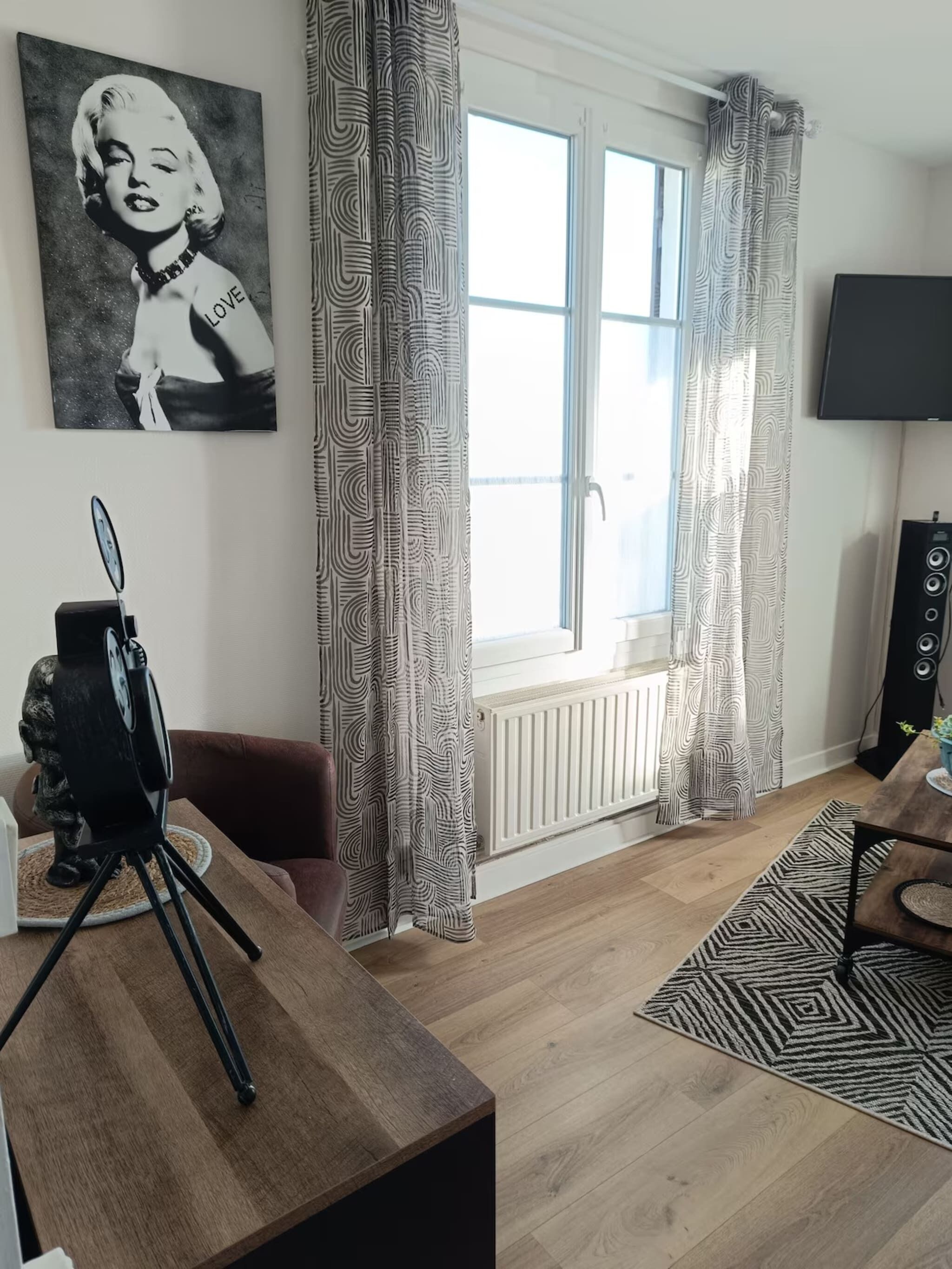 Een moderne woonkamer met een groot raam, patroon gordijnen, een statief en een ingelijste foto aan de muur.