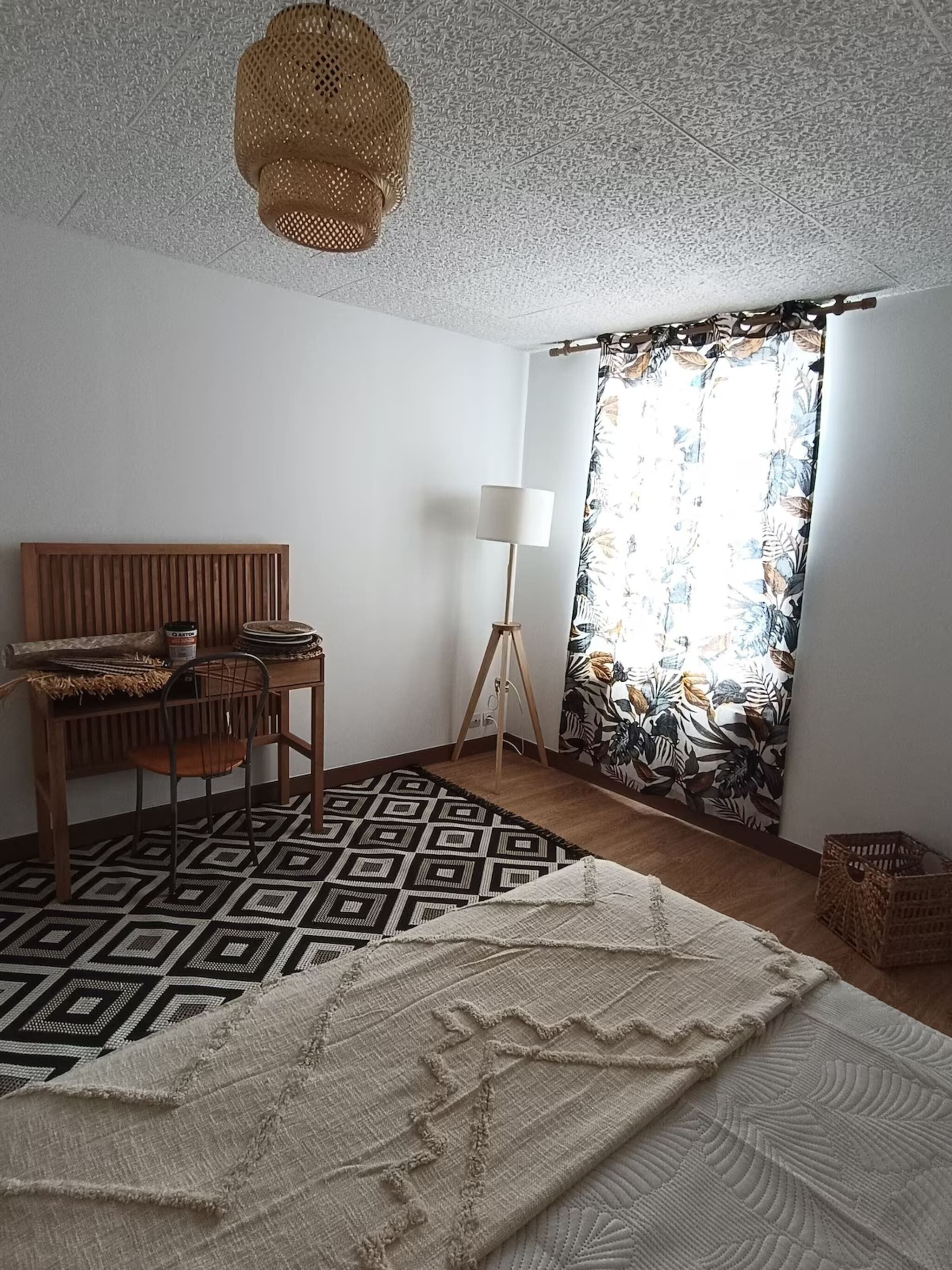 Una camera da letto con un letto, una testiera in legno, un tavolino con una lampada, un tappeto decorato e una finestra con tende floreali.