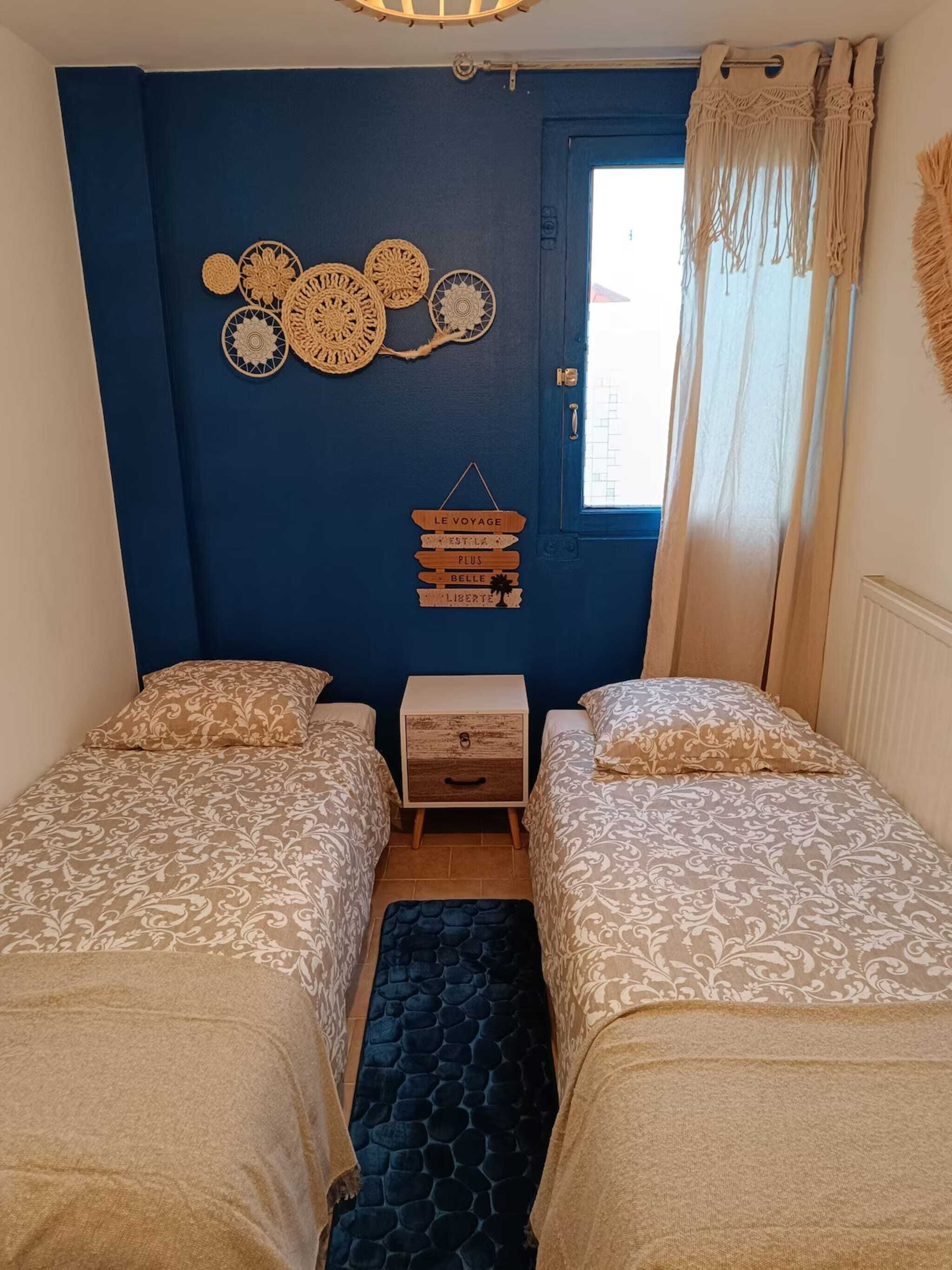 Una habitación con dos camas, una mesita de noche de madera, un adorno en la pared con un letrero y una ventana con cortinas.