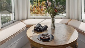 A round wooden table with a vase of flowers and a pair of binoculars on it, set in a corner with a view of a lake and greenery. - 3 bedroom accommodation in St Mawes (St Mawes)