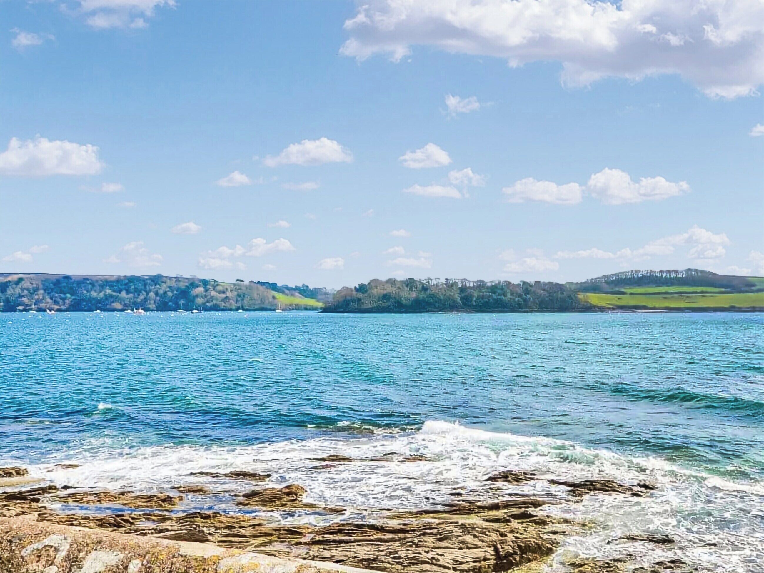 A coastal landscape with a rocky shoreline, calm water, and distant hills under a clear sky.