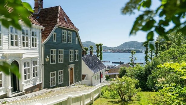 Une maison traditionnelle à deux étages, avec une façade blanche et verte, un toit recouvert de tuiles rouges et une vue sur un plan d’eau et des collines en arrière-plan.