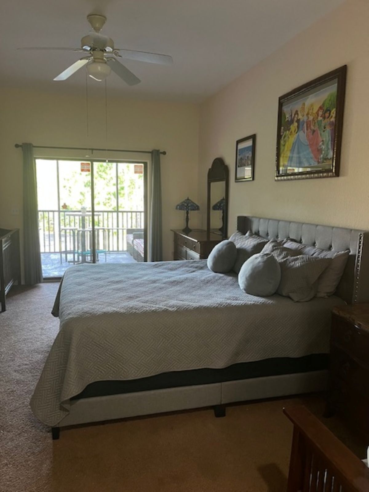 A bedroom with a large bed, a ceiling fan, a window with a view, a dresser, and a painting on the wall.