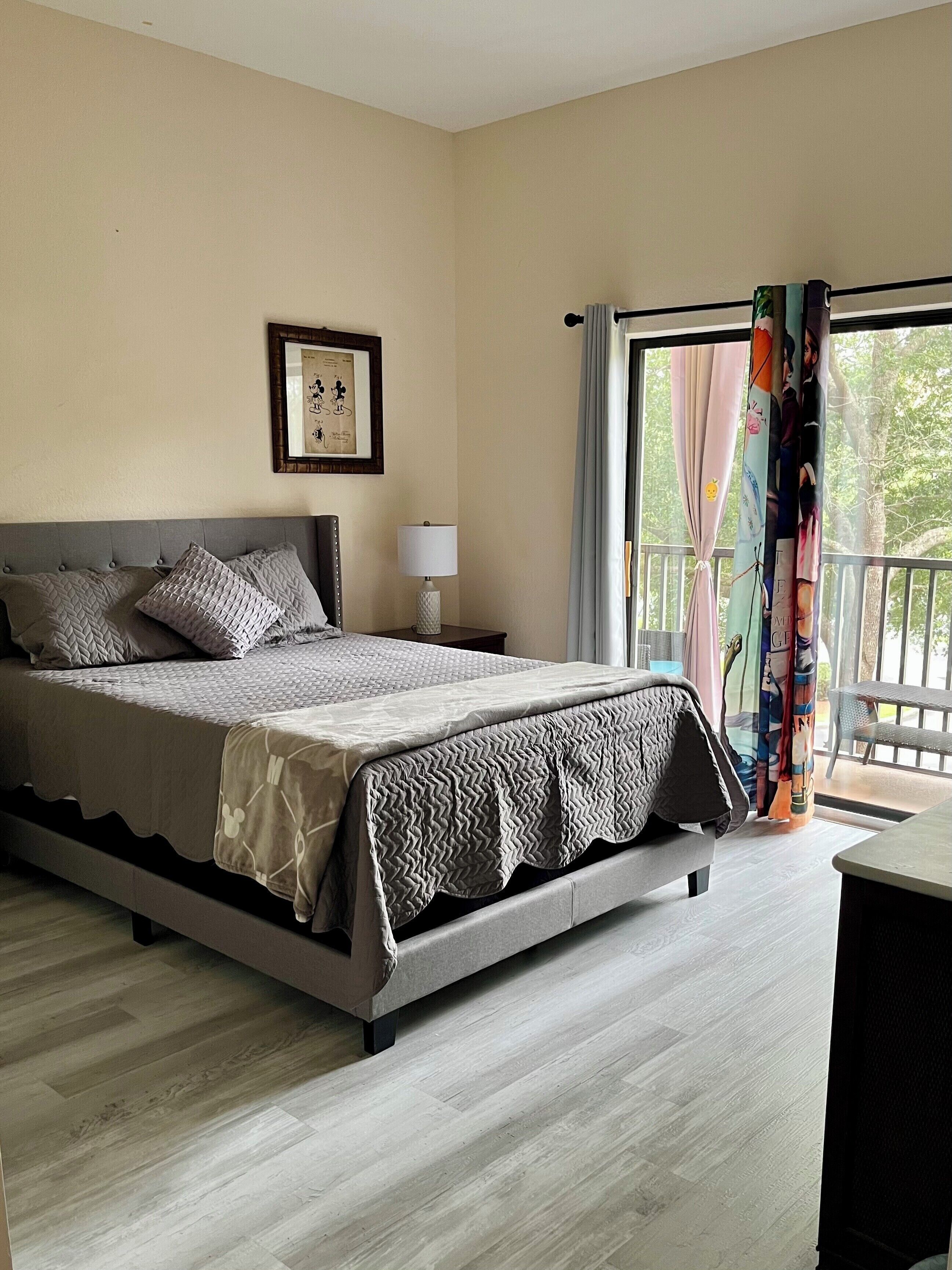 A bedroom with a bed, a nightstand, a lamp, a framed picture, and a balcony with curtains.