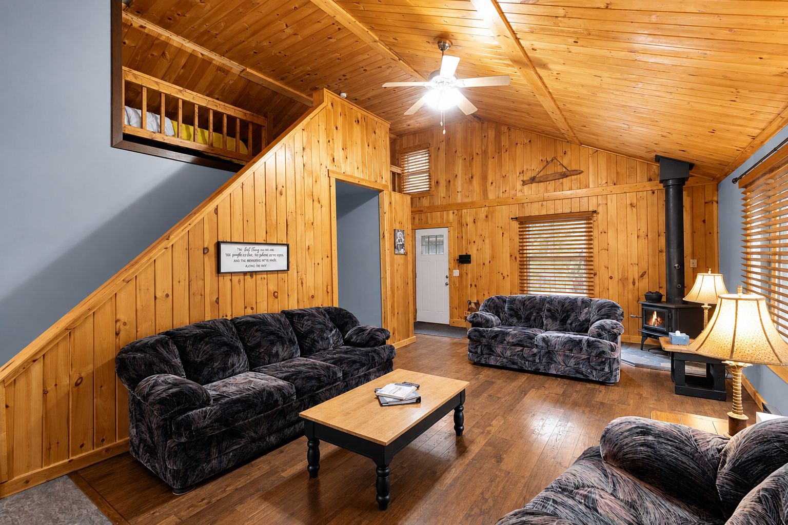 A cozy cabin interior with a wooden staircase, two patterned sofas, a wooden coffee table, a fireplace, and a ceiling fan.