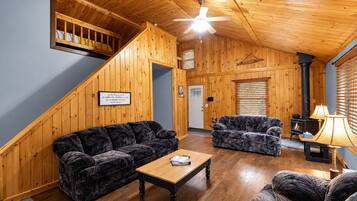 A cozy cabin interior with a wooden staircase, two patterned sofas, a wooden coffee table, a fireplace, and a ceiling fan.