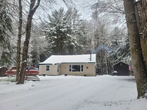 A snow-covered house with a chimney, surrounded by trees and a wooden shed.
