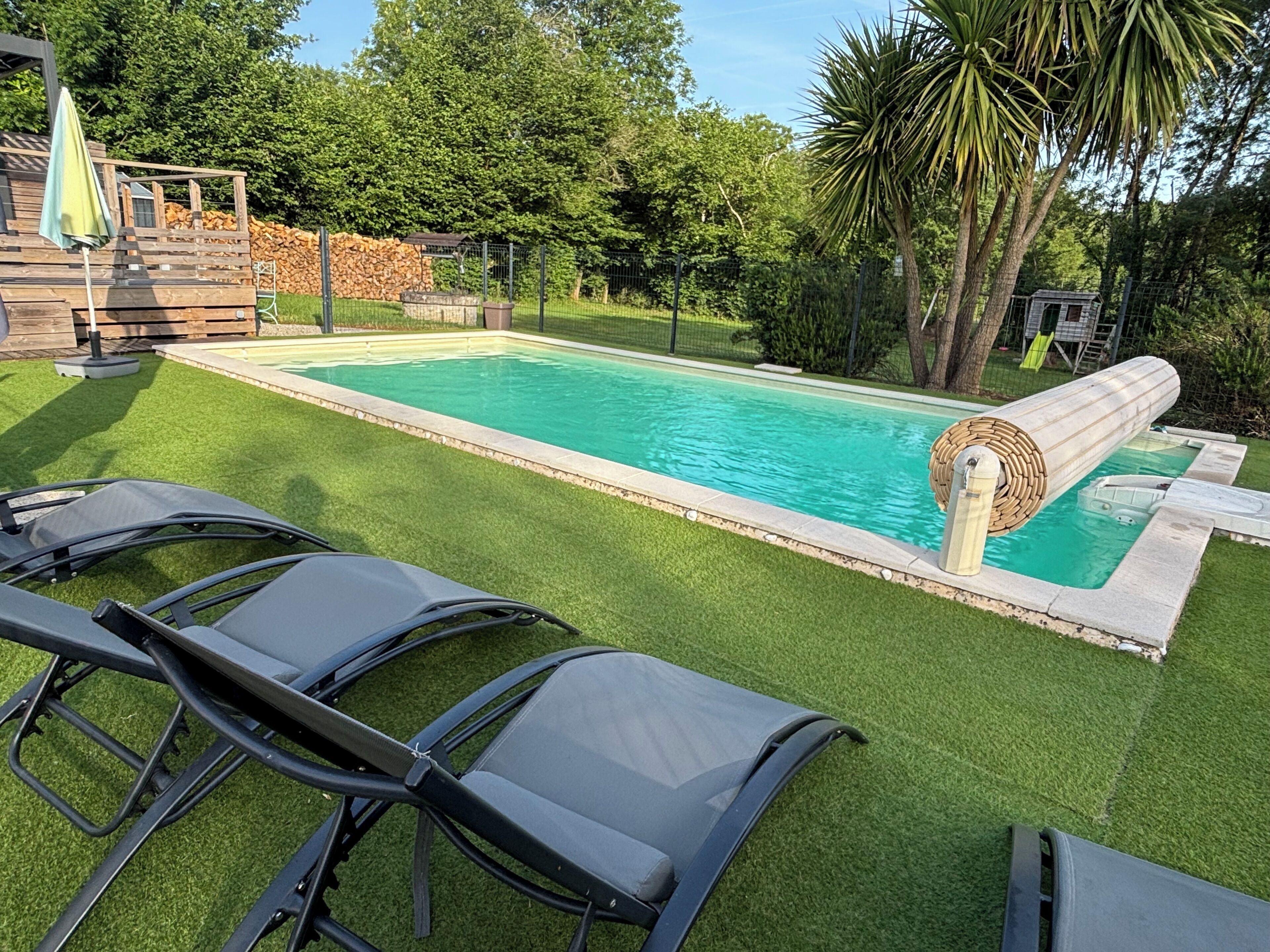 A swimming pool with a water slide, surrounded by artificial grass, and a wooden deck with chairs.