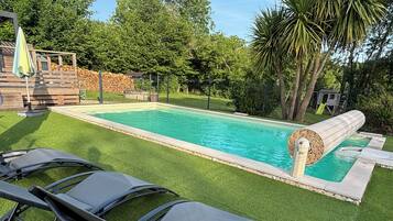 A swimming pool with a water slide, surrounded by artificial grass, and a wooden deck with chairs.
