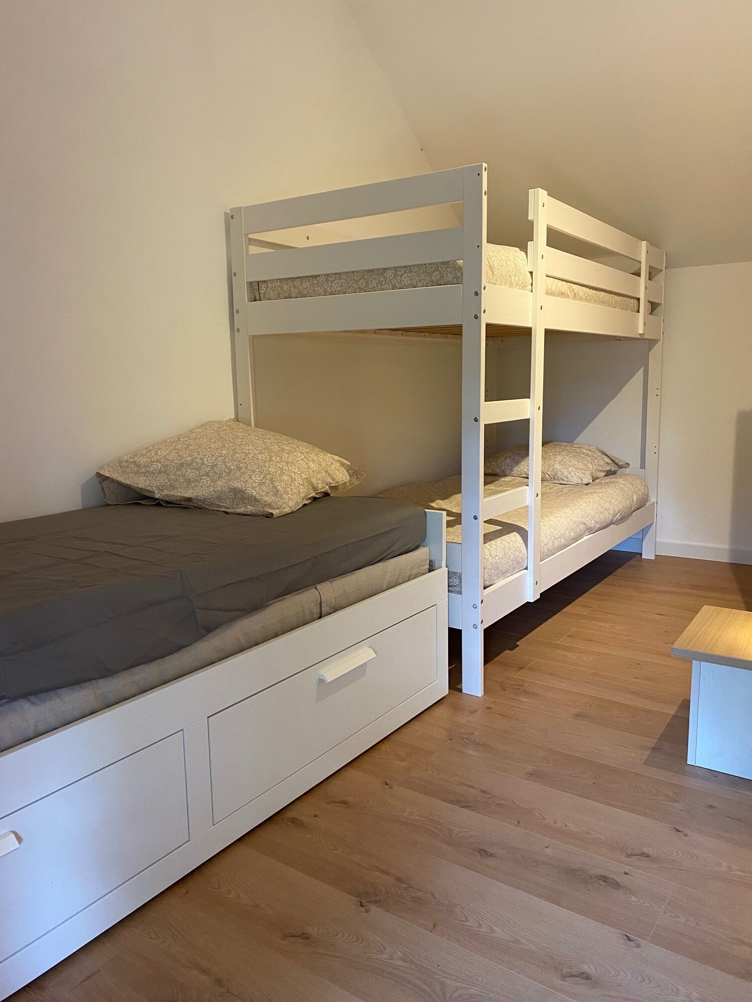 Un letto a castello con un letto inferiore e un letto singolo, entrambi con biancheria a motivi. Sotto il letto inferiore, un piccolo sgabello di legno e un mobiletto contenitore bianco.