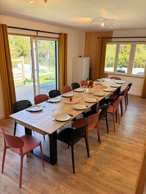 A dining area with a long table set for a meal, surrounded by chairs, and large windows offering a view of the outdoors.