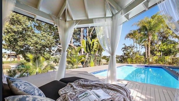 Un área de descanso junto a la piscina con mobiliario cubierto, piscina y exuberante vegetación tropical.