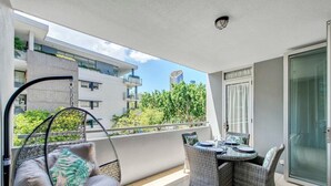A balcony with wicker furniture, a hanging chair, and a view of neighboring buildings and greenery.