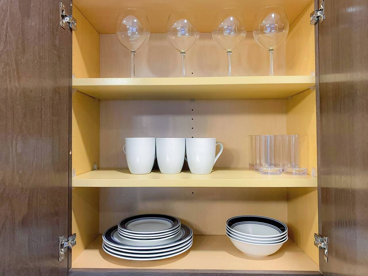 A cabinet with three shelves. The top shelf holds wine glasses, the middle shelf has white cups and glasses, and the bottom shelf contains stacked plates and bowls.