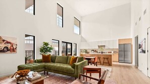 A spacious living area with a green sectional sofa, a glass coffee table, and a wooden chair. A kitchen with a stainless steel refrigerator and wooden cabinets is visible in the background. - NashVybe PH Condo| Broadway & Gulch| Pool, Parking (Nashville)