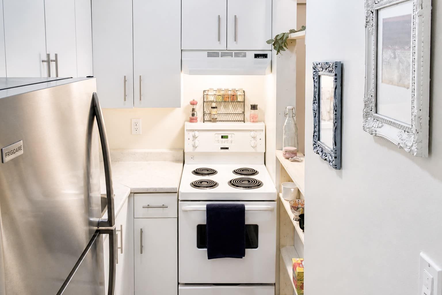 Una cocina compacta con refrigerador de acero inoxidable, estufa blanca y gabinetes blancos, además de una toalla negra.