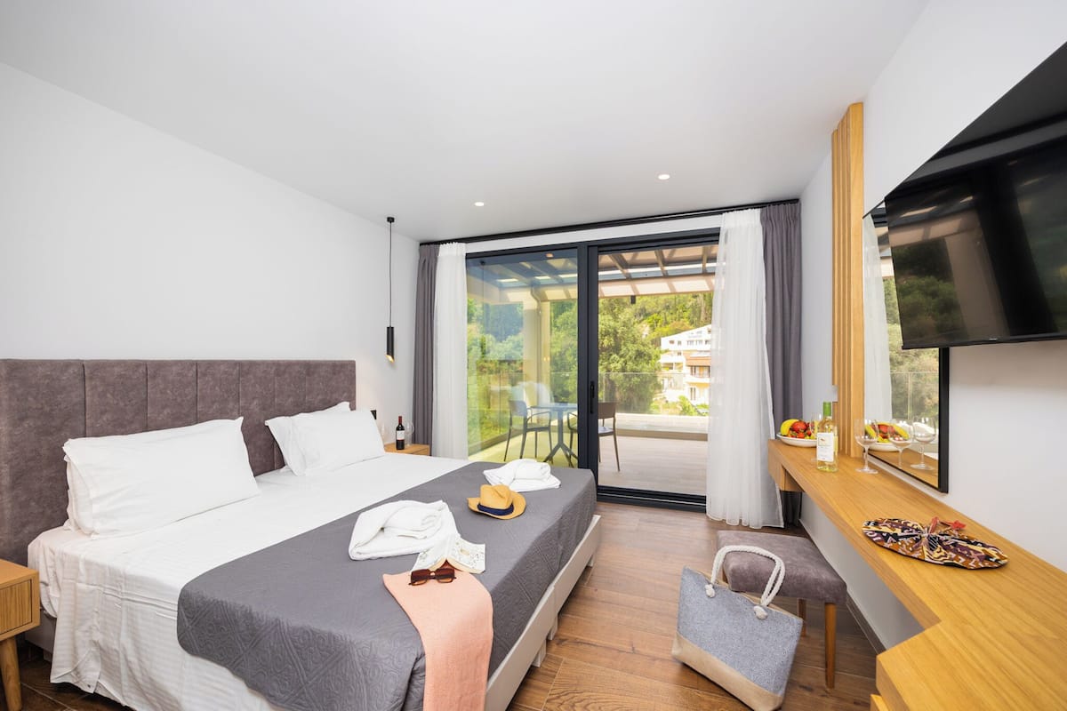 A modern hotel room with a large bed, a flat-screen TV, a balcony with a view of greenery, and a wooden side table with a bottle and fruit.