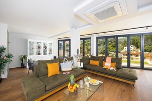 Una sala de estar moderna con un sofá grande, una mesa de centro de vidrio con fruta y bebidas, y vistas a un paisaje verde a través de amplios ventanales.