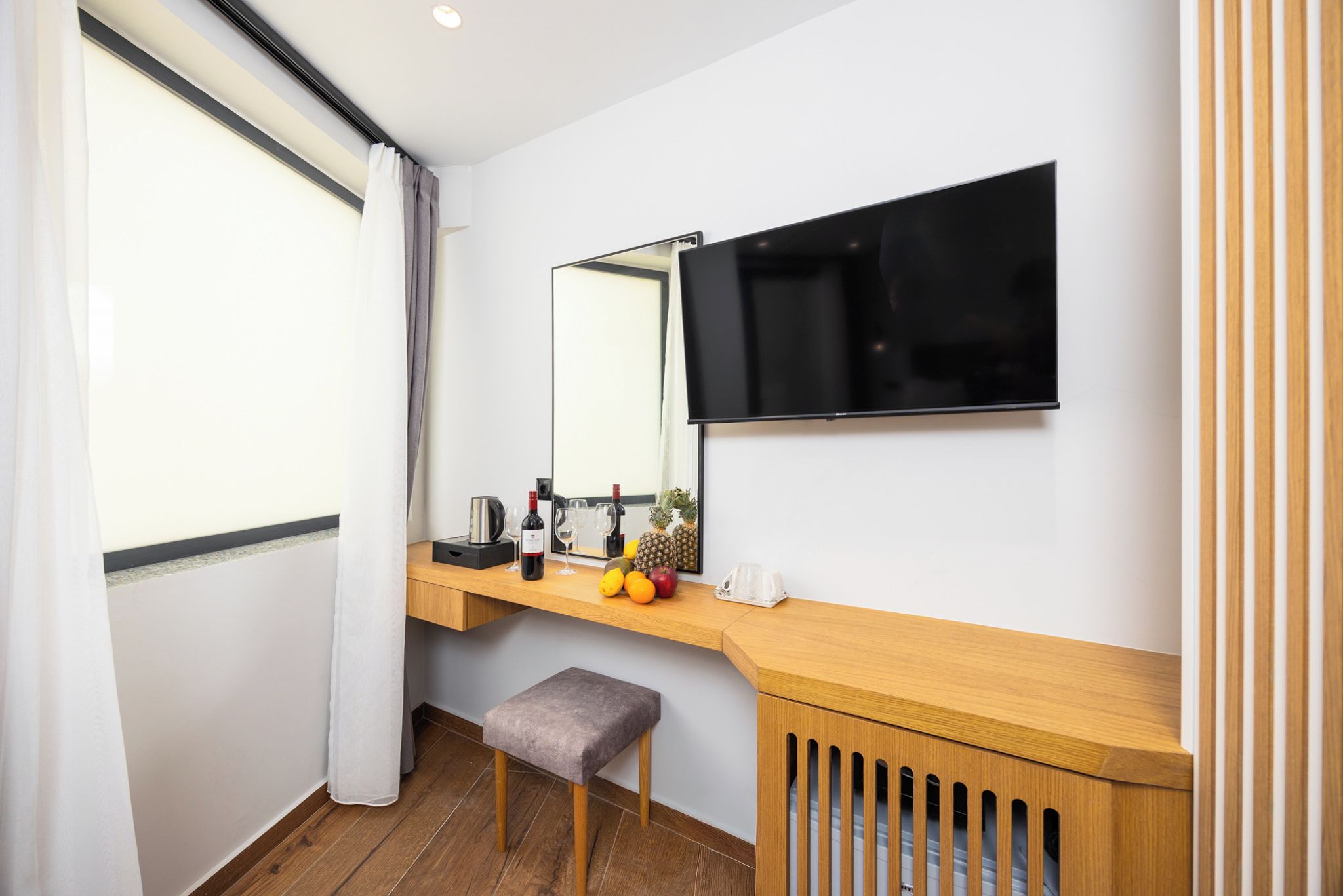 A modern hotel room with a wooden vanity, a flat-screen TV mounted on the wall, a small stool, and a window with curtains.