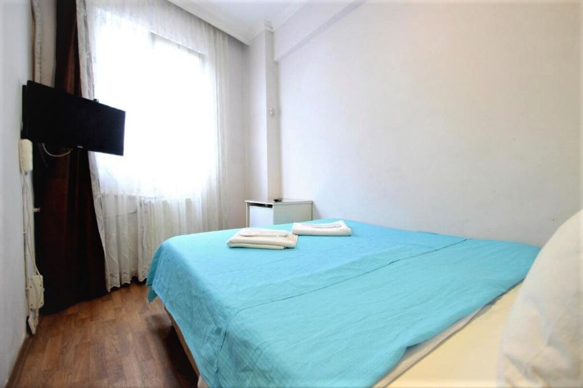 A small, clean room with a single bed, a bedside table, and a window with curtains.